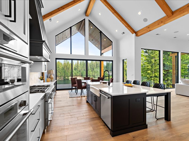 Kitchen with a dark island, vaulted ceiling beams, appliances, and large windows.