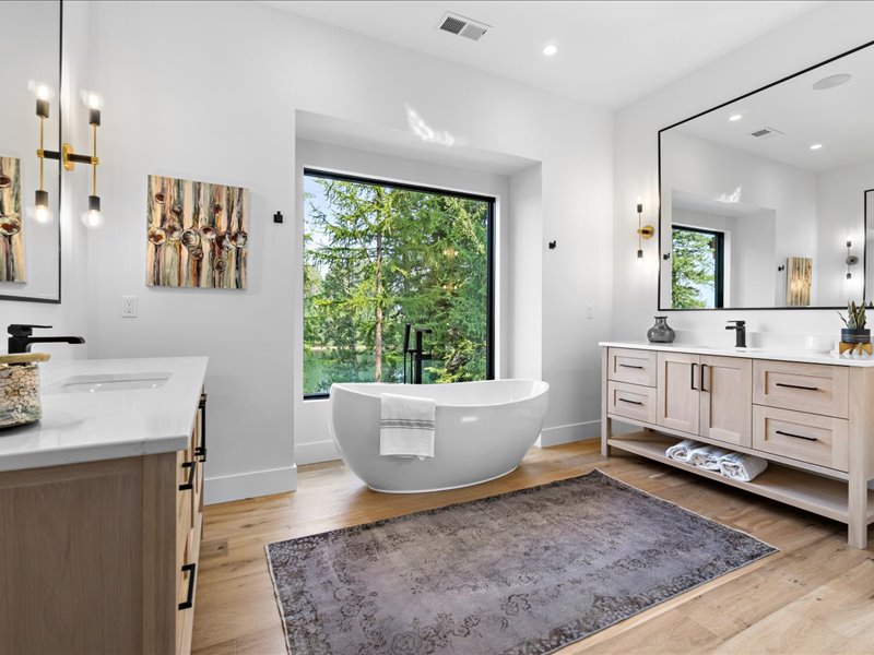 Bathroom with a freestanding tub, double vanity, brown plank-pattern flooring, and a large window facing trees.